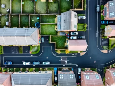 A suburban street in England, shown directly from above, with lush, green yards and driveways with cars parked in front.