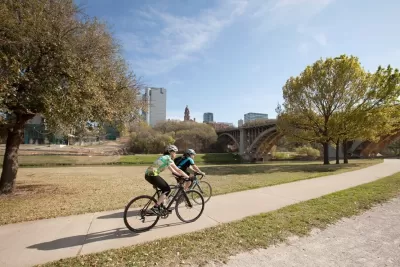Two cyclists on a paved bike trail through short grass with Dallas buildings in background