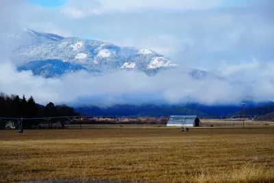 A long farmhouse is visible in winter agricultural fields, Snow-covered mountains loom above low clouds in the background of the photo.