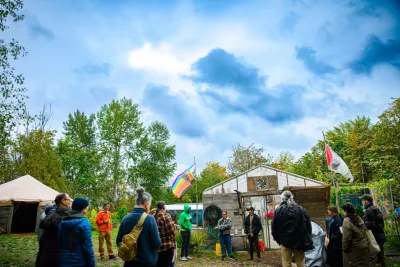 A group of people circle around in front of the Heron’s Nest greenhouse entrance. Two flags ever in the wind as a cloudy sky hovers over the volunteers of the space.