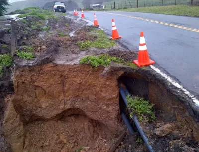 Eroded cliffside and exposed utilities along asphalt Highway 1 in California with orange traffic cones lining side of road.