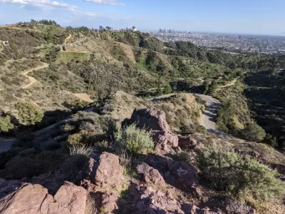 The Griffith Observatory is perched on a hill above Los Angeles, with the city's downtown shown int he distant background. Griffith Park's rolling hills and winding roads are shown in the foreground.