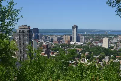 The downtown high rises of Hamilton, Ontario are surrounded by greenery and the blue of Lake Ontario on a sunny day.