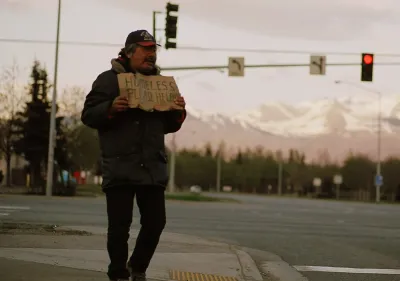 Unhoused man holds up cardboard sign on street corner in Anchorage, Alaska with snowy mountains in background.