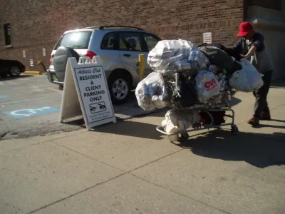 Unhoused man wearing a red bucket hat pushing a shopping cart full of plastic bags full of empty cans.