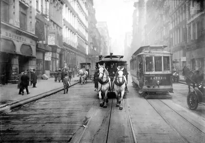 A black-and-white photo of a team of white horses pulling a trolley car along a track on a New York City Street, facing the camera.