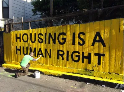 Man in green shirt painting yellow and black "Housing is a human right" mural on fence.