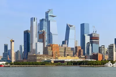 A large group of glass covered skyscrapers rises above the Hudson River in Manhattan, with several new buildings under construction visible too.
