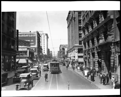 Black-and-white 1924 photo of a street in Los Angeles with a street car, cars driving in the other lane over street car tracks and pedestrians walking on sidewalk and middle of street.