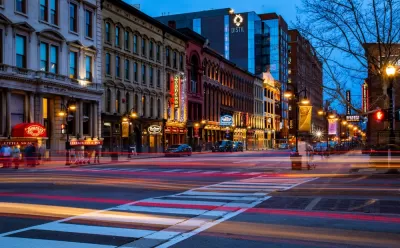 A Downtown Louisville street at night, with the lights of passing shown with a blurred effect.
