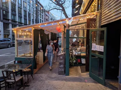 Person wearing mask walking through temporary outdoor dining setup lined with bistro lights at dusk in New York City.