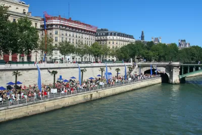 A large crowd gathers on the banks of the River Seine on a sunny day in Paris.