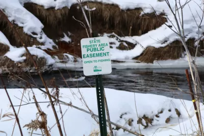 A sign reads “State Fishery Area Public Easement Open to Fishing.” In the background is a stream surrounded by snow on the ground in a natural setting.