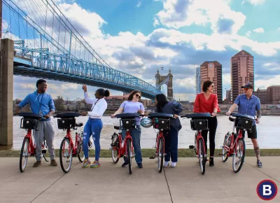 Six people posing with Red Bike bike share bikes with bridge and river in background.