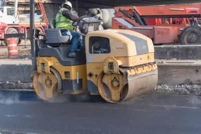A man operates a yellow roller to smooth out fresh pavement on a road.