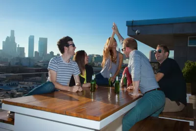 A group of friends laughs and celebrates with beers on a rooftop with Downtown Los Angeles in the background.