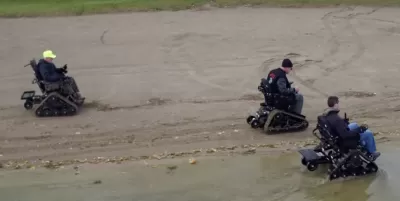 Aerial view of three men using track chair mobility devices on lake beach in Michigan state park.