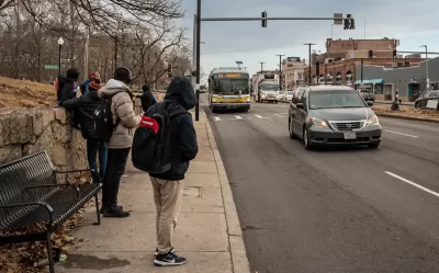 People in hoodies and jackets waiting for oncoming bus at sidewalk bus stop in Boston, Massachusetts.