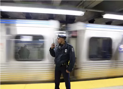 Transit police officer standing on subway platform with blurred train passing behind him.