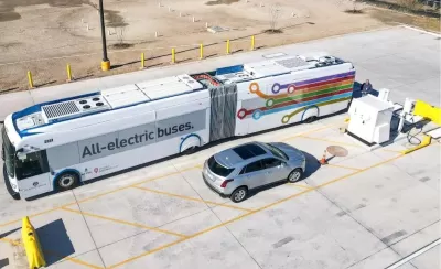 Aerial view of electric bus parked next to silver SUV in parking lot in Austin, Texas. Bus says "All eelctric buses" on side.