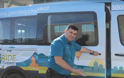 Man in teal shirt opening door to white microtransit shuttle with cactus graphics and making inviting gesture toward the camera.