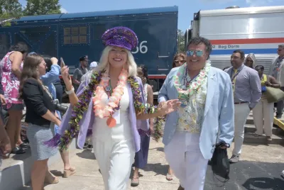 Blonde woman in sequined purple hat walking away from Amtrak train smiling at camera with other people behind her.