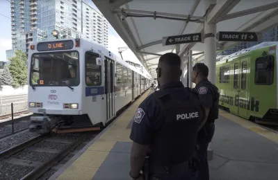 Two transit police officers standing on outdoor train platform in Denver with white light rail train approaching.