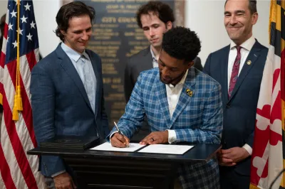Mayor Brandon M. Scott of Baltimore wearing blue checkered suit signing a bill with three men in suits standing behind him at podium.
