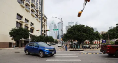 Blue SUV turning left in intersection in Austin, Texas with pedestrians waiting to cross and Lime scooters lined up on sidewalk.