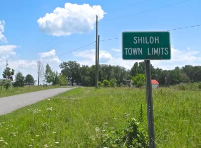 Green sign with white text 'Shiloh Town Limits' against green grassy field with small road on left.