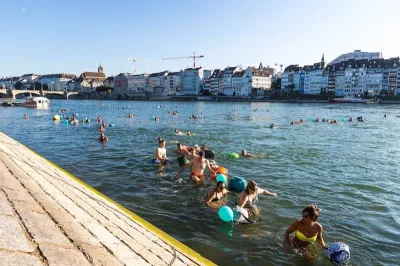 People swimming in Rhine River on a sunny day with low-rise, dense buildings along waterfront in background.