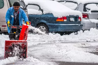 A man pushes a snow blower to clear the ground of an outdoor parking lot filled with cars.