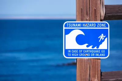 A sign, with the blue ocean int he background, reads "Tsunami Hazard Zone: In Case of Earthquake Go to Higher Ground or Inland"