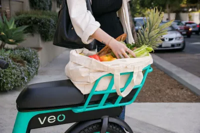 Close-up on back basket of teal and black Veo bike share bike with woman's hands reaching into canvas bag full of produce.