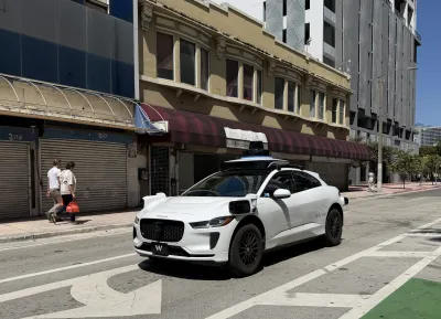 White Waymo robotaxi on street in Miami, Florida with two men walking on sidewalk in background.