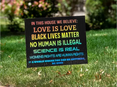 A rainbow colored sign planted in the front yard of a typical suburban home reads, "In this house we believe love is love, black lives matter, no human is illegal, science is real, women's rights are human rights. In a world where you can be anything, be kind."