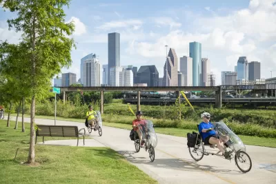 Three men riding recumbent bikes on paved greenway next to bayou with Houston skyline in background.