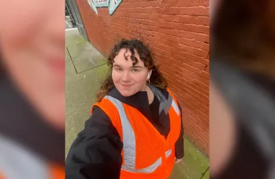 Woman taking selfie while wearing orange safety vest outdoors in front of red brick wall on field visit.
