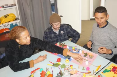 Three people gather around a table to discuss maps made of toys and found objects.