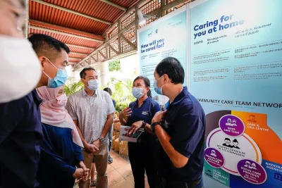 Two people in masks stand in front of a "caring for you at home" sign. They speak to people in medical masks.