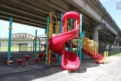 Red, yellow, and green plastic playground equipment in small park under Claiborne Expressway overpass in New Orleans, Louisiana.