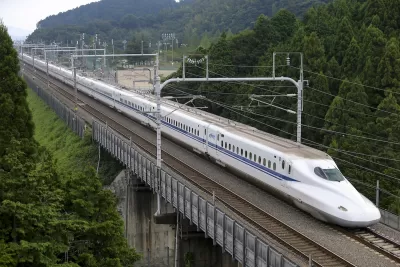 Shinkansen high-speed rail train traveling across bridge with green trees on either side.