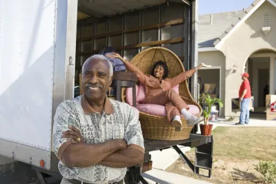 African American senior couple smiling in front of a moving truck in front of a single-family home.
