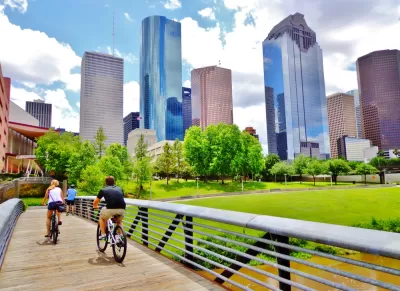 Two cyclists on a wooden bike bridge across a bayou with downtown Houston skyline in background