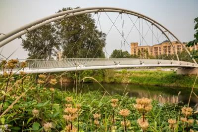 View of pedestrian suspension bridge over waterway surrounded by green grass with small apartment buildings in background