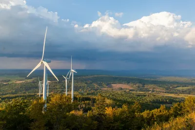 A cluster of wind turbines beneath a stormy sky, above a forest where the leaves are beginning to change colors.