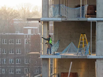 Construction worker in safety gear stands in an unfinished concrete high-rise building with older brick buildings visible in background.