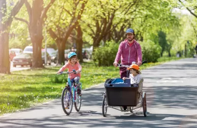 Father riding cargo bike with one child in cargo basket and one child riding bike alongside on sunny tree-lined path