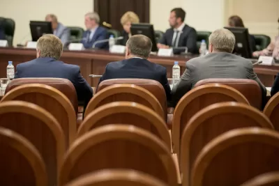 View from back of three men in suits sitting in a large room with leather chairs facing dais