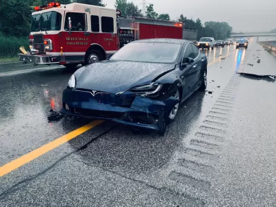 Dark blue Tesla with crumpled front bumper on rainy highway with red fire truck in background